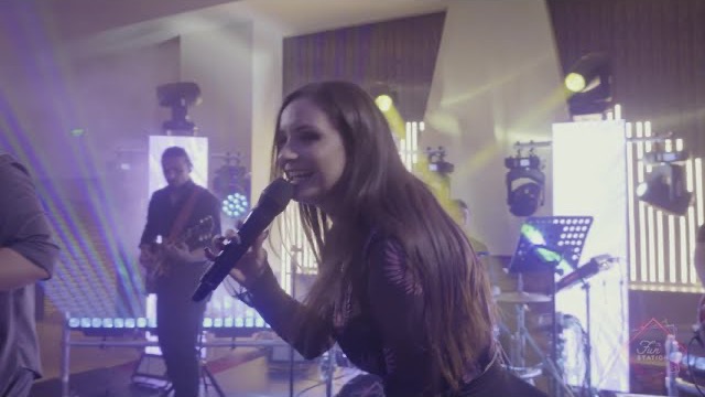 Solista trupei cântă zâmbind la microfon, cu chitaristul și luminile scenei în spate