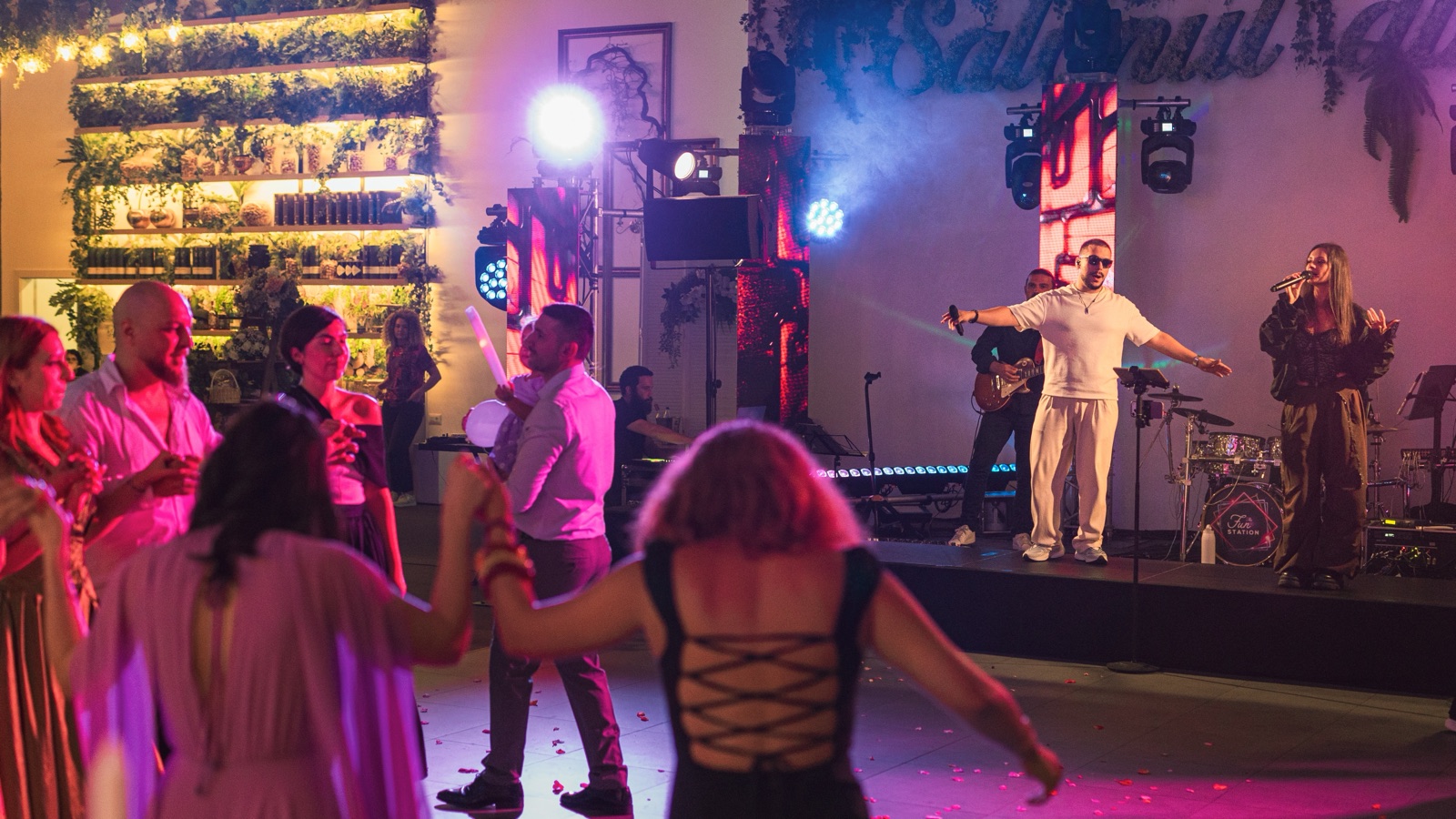 Trupa pe scenă, cu solistul în alb cu brațele deschise și solista la microfon, iar în față invitații dansează în lumină roz