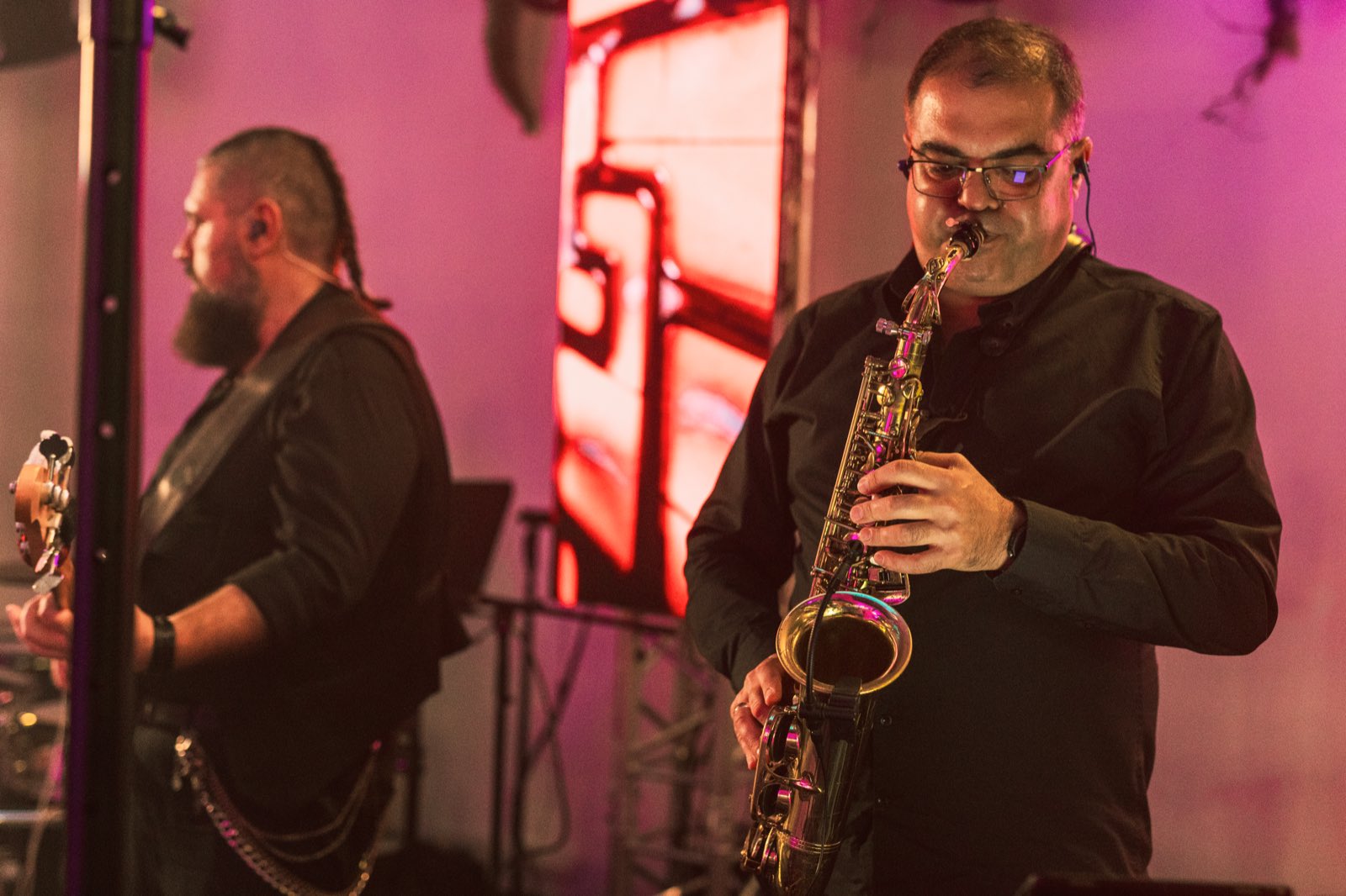 Saxofonistul trupei cântând la saxofon pe scenă, în lumină roz, cu basistul în stânga și un ecran LED în spate