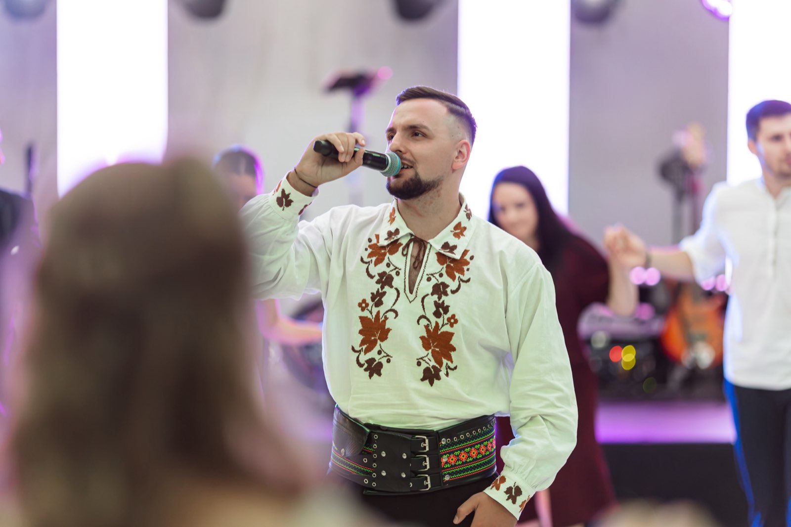 Solistul de muzică populară, în cămașă tradițională brodată, cântând la microfon printre invitați