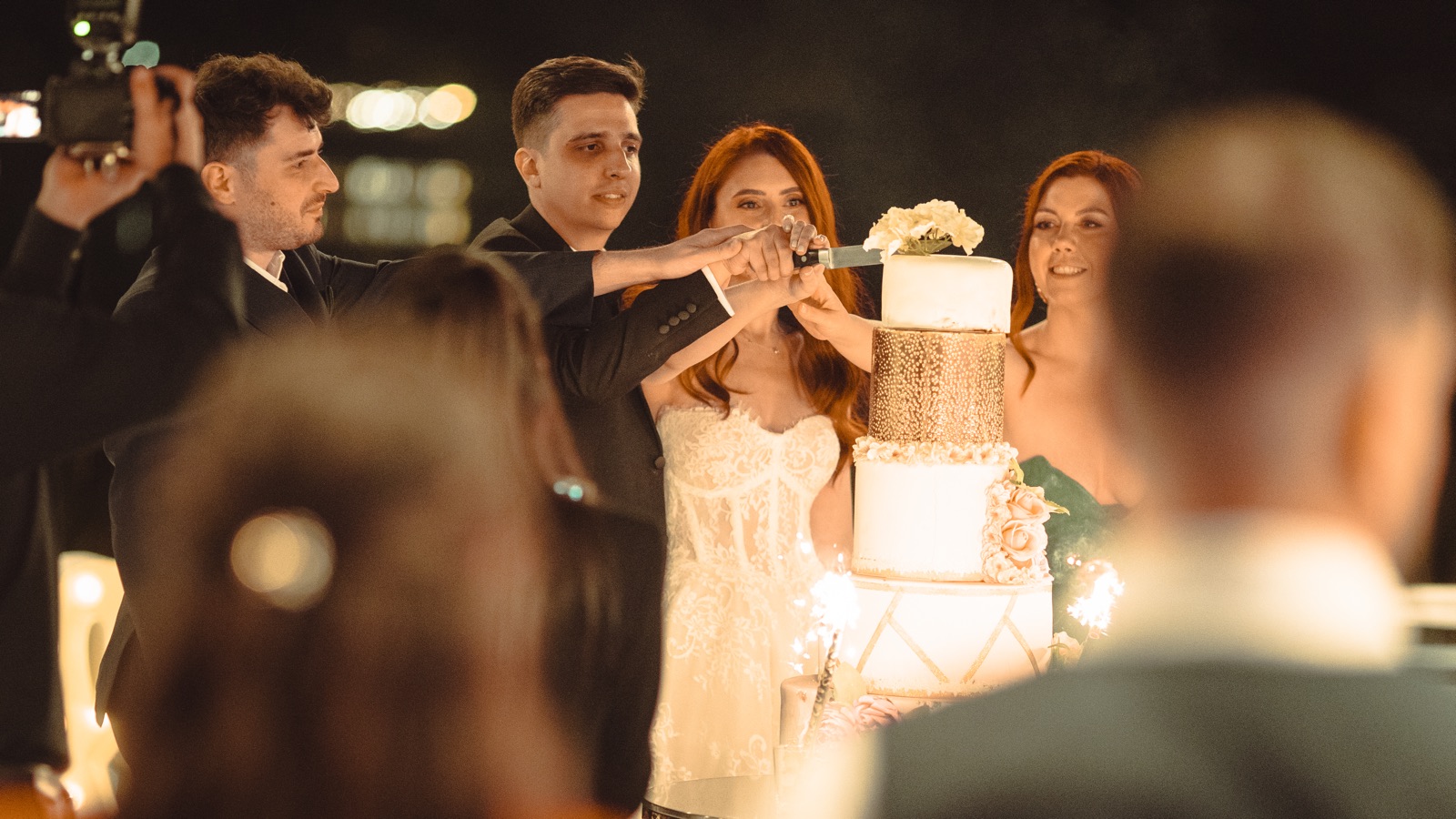 Mirii taie tortul afară, seara, cu artificii de tort aprinse și cameramanul filmând din stânga