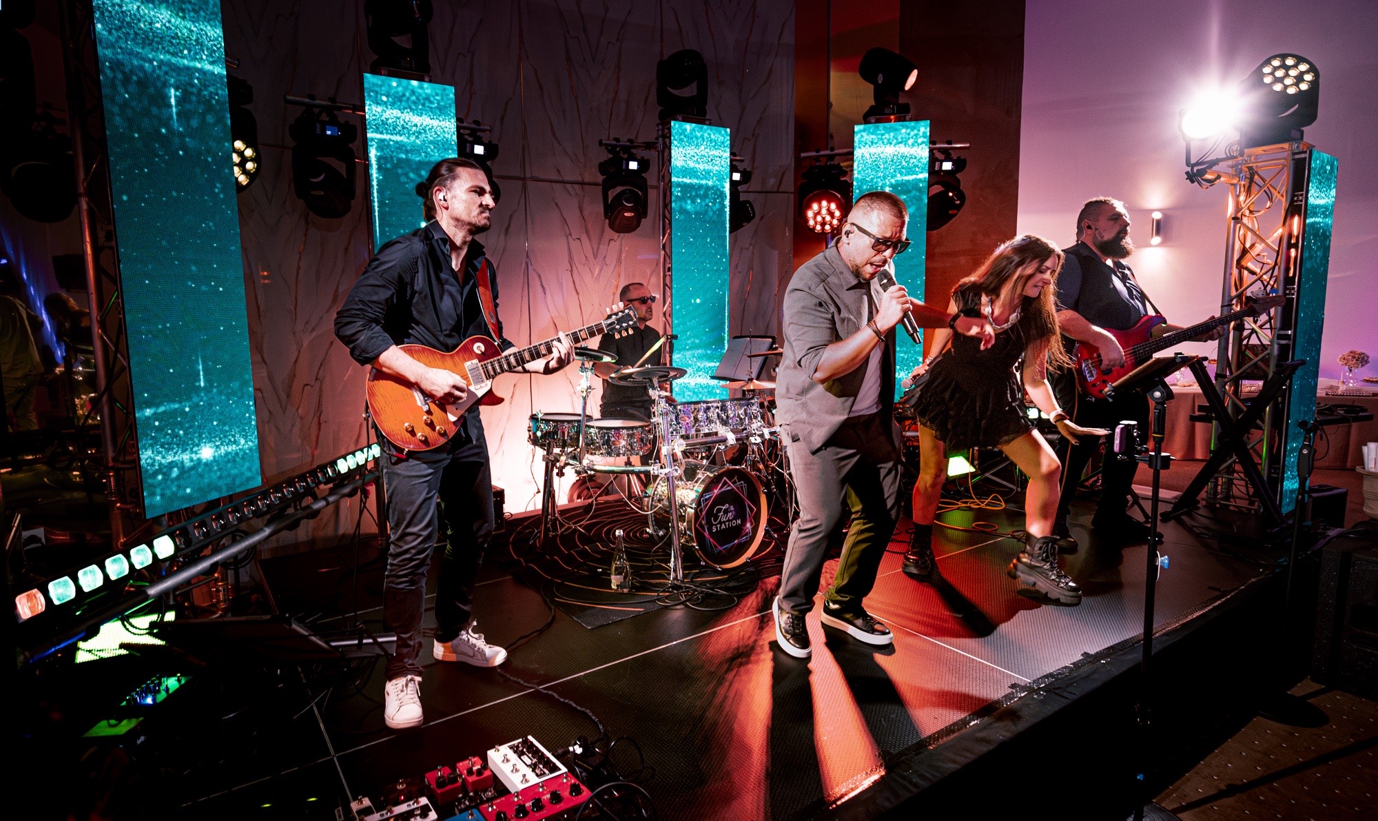 Trupa The Fun Station Band pe scenă în timpul unui eveniment: chitară, tobe, solist și solistă, bas, cu ecrane LED în spate