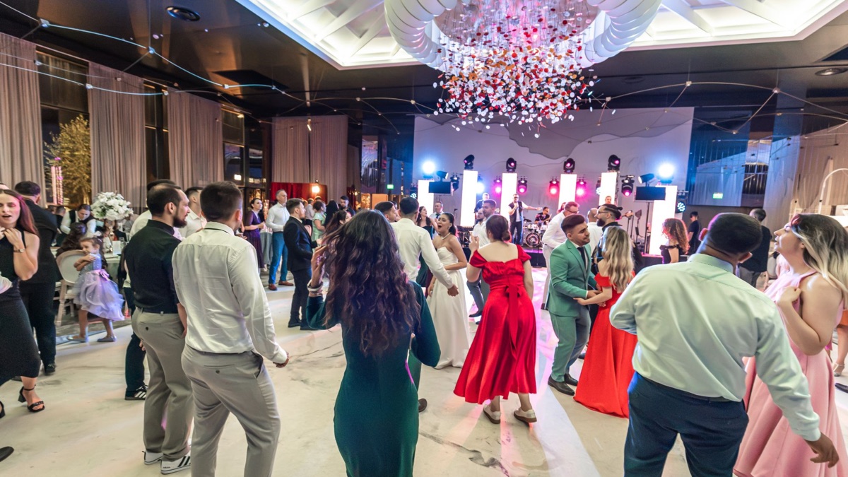 Ringul plin de invitați care dansează, cu trupa cântând pe scenă între stâlpii LED, sub candelabrul sălii