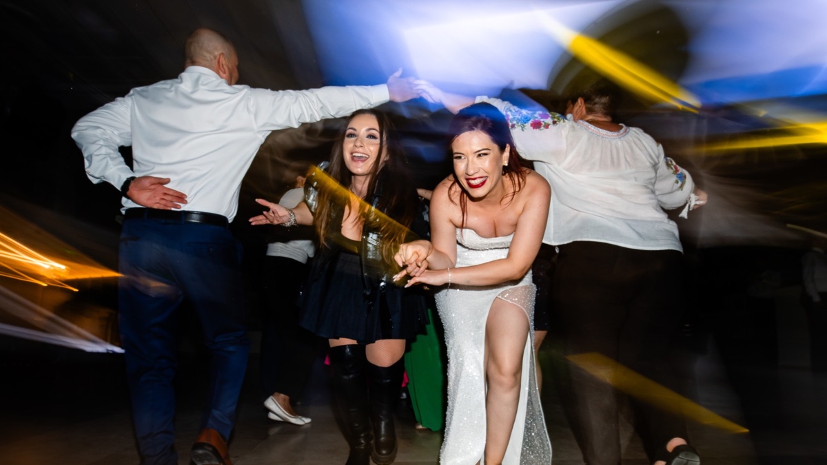 Mireasa și o invitată râzând, trecând pe sub brațele ridicate ale celor care dansează